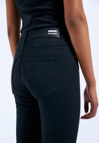Black denim jeans with a fitted silhouette, featuring back pockets and a branded label at the waistband. Smooth texture, classic design.