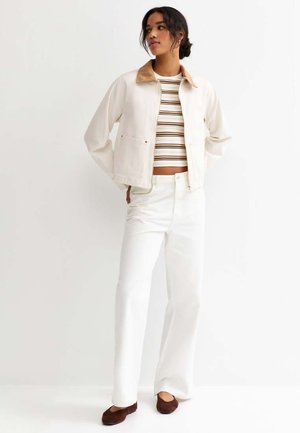 Kobieta stojąca, ubrana w kremową kurtkę z beżowym kołnierzem, prążkowany krótki top, białe spodnie o szerokich nogawkach i brązowe baleriny na jednolitym tle.