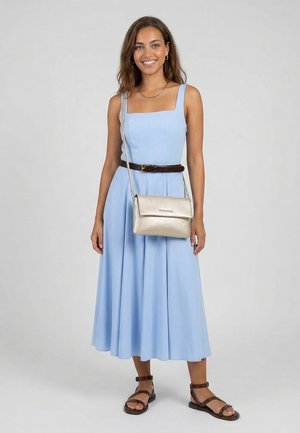 Smiling woman wearing light blue sleeveless midi dress with brown belt, dark brown sandals, gold crossbody bag, and gold necklace, standing.