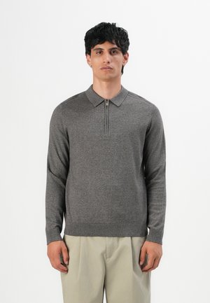 Man wearing a gray long-sleeve sweater with a zipper collar and beige pants, standing against a plain white background.
