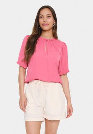 Blouse rose à manches bouffantes courtes, avec un col en volants et un détail en forme de clef. Associée à un short beige clair à taille élastique.