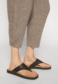 Pieds portant des sandales en cuir noir texturé avec des semelles couleur tabac, associées à un pantalon en maille beige raccourci.