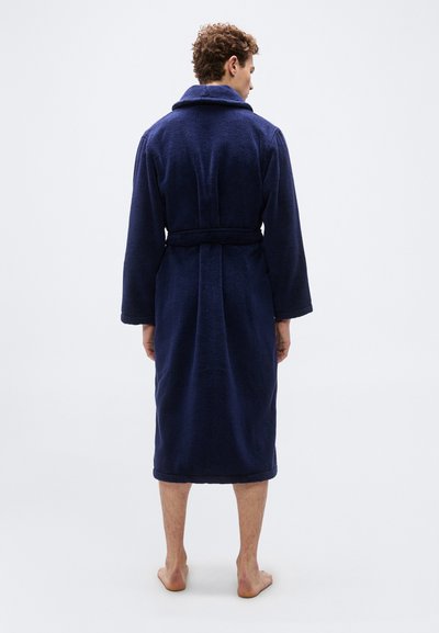 Άνδρας με μπλε ρόμπα μέχρι τα γόνατα με ζώνη, όρθιος ξυπόλυτος με την πλάτη γυρισμένη σε απλό λευκό φόντο.