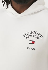 Uomo di colore con dreadlock corti che indossa un hoodie bianco con il logo "HILFIGER NEW YORK" e un emblema di bandiera rossa, bianca e nera sul petto.
