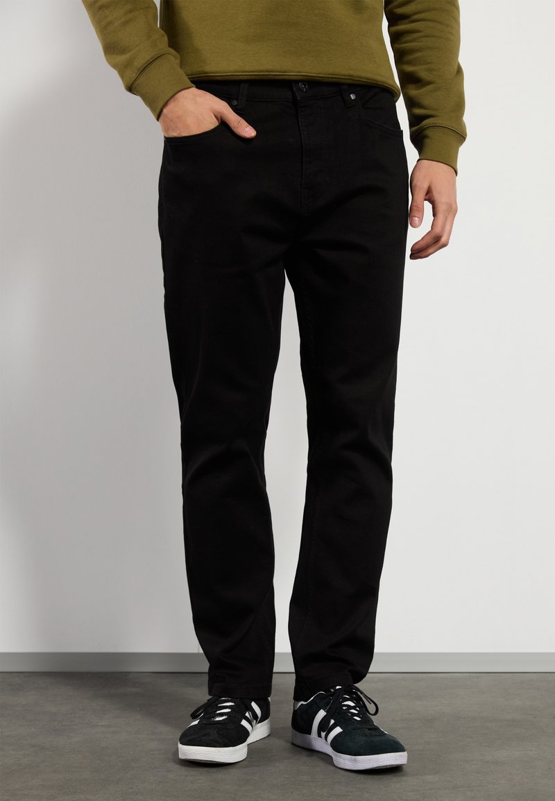 Jeans de mezclilla negra con un diseño de pierna recta, que cuentan con bolsillos y cierre de botón. Combinados con zapatillas negras y blancas.