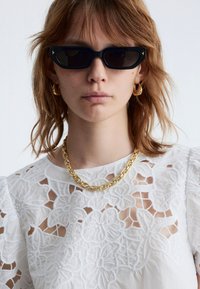 Black rectangular sunglasses, gold chain necklace, and floral embroidered white top. Features a textured design with cut-out details.