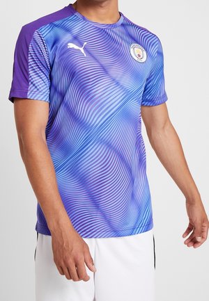 Homme portant un maillot de sport Manchester City à manches courtes bleu et violet avec un motif ondulé et le logo Puma, associé à un short blanc.