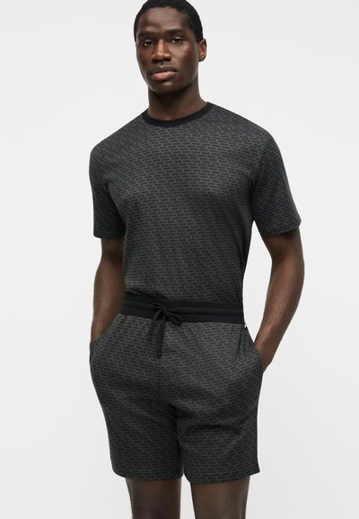 Homme portant une chemise à manches courtes gris foncé et un short assorti avec un motif de texte répétitif subtil, mains dans les poches, fond neutre.