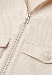 Close-up of beige textured fabric jacket with silver zipper and buttoned flap pocket.