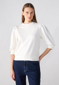 White pullover with a high neckline and puffed sleeves. Smooth texture, fitted at the waist, paired with dark blue jeans.