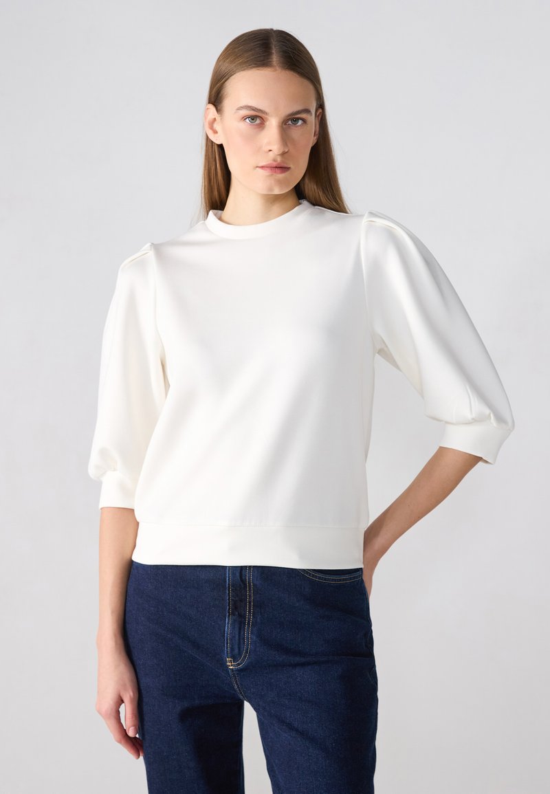 White pullover with a high neckline and puffed sleeves. Smooth texture, fitted at the waist, paired with dark blue jeans.