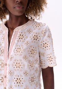 Witte blouse met beige bloemenborduurwerk, ronde halslijn, korte mouwen en decoratieve knopen. Gestructureerde stof met doorschijnende accenten.