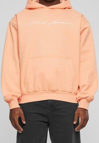 Persona che indossa un hoodie pesca con la scritta bianca "Karl Kani" sul petto e jeans neri, in piedi contro uno sfondo grigio uniforme.