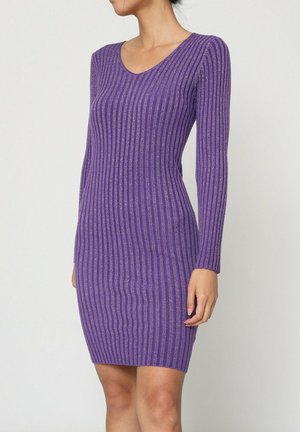 Femme portant une robe moulante à manches longues côtelée violette, debout devant un fond clair uni.