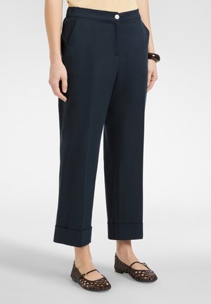 Mujer con pantalones azul marino con puños y zapatos planos tejidos de color marrón oscuro, de pie con las manos relajadas a los lados.