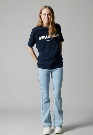 Giovane donna sorridente, indossa una t-shirt blu navy con la scritta "WHILED AWAY", jeans azzurri e sneaker bianche, in piedi davanti a uno sfondo grigio.