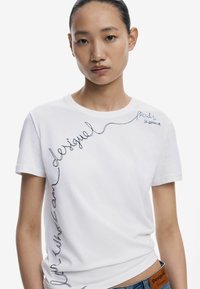 Valge puuvillane t-särk lühikeste varrukatega, millel on must tikand, kus on kirjas "desigual" ja "proud to be different". Lihtne ja vabaltaja disain.