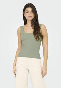 Top de tirantes acanalado en verde oliva con bordes en forma de ola, presenta tirantes gruesos y un diseño ajustado. Combinado con pantalones de pierna ancha en beige claro.