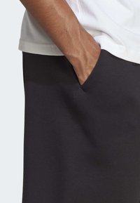 Mustat housut, joissa on sivutasku, valmistettu pehmeästä kankaasta, jossa on sileä pinta. Yllä oleva paita on valkoinen ja osittain näkyvissä.