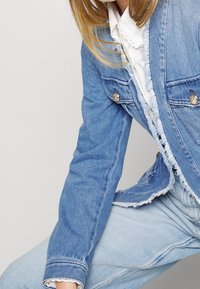 Chaqueta de mezclilla azul clara con bordes deshilachados, dos bolsillos en el pecho y botones a presión plateados. Llevada sobre una camiseta blanca con cuello. Diseño informal.