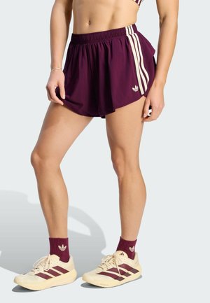Femme portant un short de sport violet avec des rayures blanches, des chaussettes assorties et des chaussures de course beige avec des rayures violettes, debout sur un fond clair.