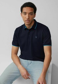Navy polo shirt with turquoise accents, short sleeves, three-button placket, and subtle logo detail. Light-wash jeans complete the look.