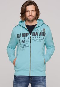 Light blue zip-up hoodie with kangaroo pockets, featuring black printed text and logo accents. Finished with a drawstring hood and ribbed cuffs.
