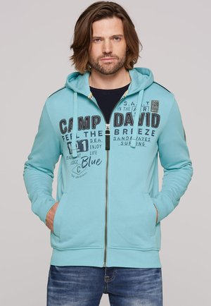 Light blue zip-up hoodie with kangaroo pockets, featuring black printed text and logo accents. Finished with a drawstring hood and ribbed cuffs.