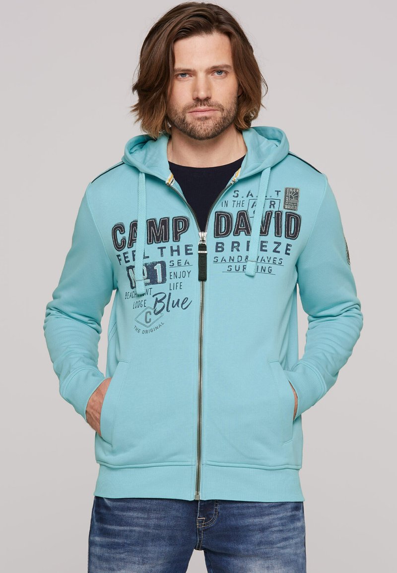 Light blue zip-up hoodie with kangaroo pockets, featuring black printed text and logo accents. Finished with a drawstring hood and ribbed cuffs.