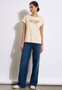 Camiseta de color crema, de manga corta, con "Las cosas buenas están por venir" impreso en cursiva roja, combinada con pantalones vaqueros de pierna ancha y zapatos blancos.