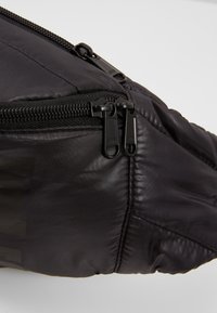 Pochette en tissu nylon noir avec deux fermetures éclair noires visibles et des détails de couture sur un fond clair.
