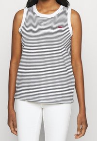 Femme portant un haut blanc sans manches avec de fines rayures noires horizontales et un pantalon blanc, arborant un petit logo rouge Levi's sur la poitrine.