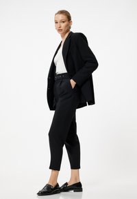 Blazer noir ajusté sur un top blanc, assorti à un pantalon noir taille haute et des mocassins noirs brillants. Design minimaliste et coupe.