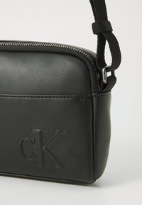 Sac bandoulière en simili cuir noir avec une finition texturée, doté d'une fermeture éclair et du logo "cK" en relief sur le devant.
