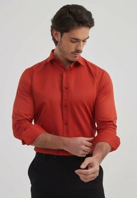 Chemise rouge à manches longues, boutonnée, avec un col structuré, une texture lisse et une coupe ajustée. Poignets retroussés et cinq boutons à l'avant.