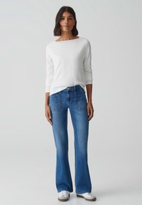 Witte lange mouwen top met een bootneck, gecombineerd met lichtblauwe flared jeans met zakdetails en een knoopsluiting, gedragen met witte sneakers.