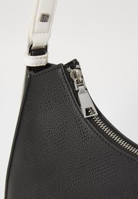 Borsa in pelle nera texture con dettagli in metallo argentato, dotata di chiusura a zip e una tracolla bianca a contrasto. Logo DKNY visibile.