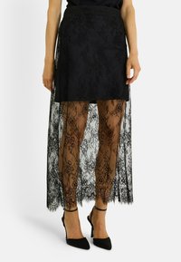 Jupe en dentelle noire avec un voile transparent, longueur genou, ornée de motifs floraux complexes. Associée à des chaussures à talons hauts noires.