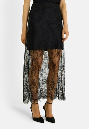 Falda de encaje negro con una superposición transparente, hasta la rodilla con patrones florales intrincados. Combinada con zapatos de tacón alto negros.