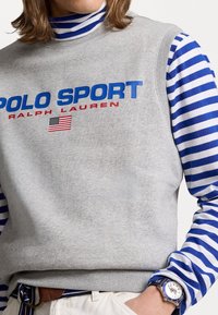 Šedá mikina s nápisem "POLO SPORT" v modré a červené, vrstvená přes modrou a bílou pruhovanou košili s dlouhým rukávem, která ukazuje hodinky na zápěstí.