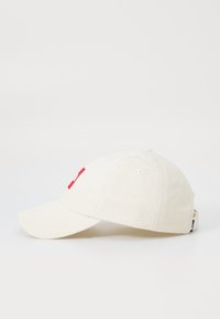 Gorra de béisbol en tela crema clara, con un parche rectangular rojo y letras blancas. Ojalillos para transpirabilidad, visera curva.