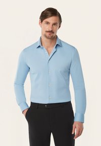 Lichtblauwe blouse met lange mouwen en een puntkraag, met een knoopsluiting aan de voorkant en een geweven stof. Gecombineerd met donkere broek.