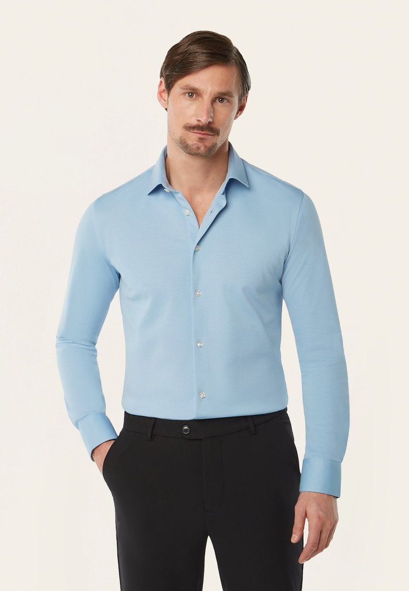 Lichtblauwe blouse met lange mouwen en een puntkraag, met een knoopsluiting aan de voorkant en een geweven stof. Gecombineerd met donkere broek.
