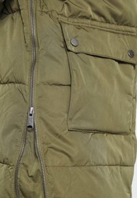 Olive green quilted jacket with exposed metal zipper and flap pocket secured by two metal snap buttons.