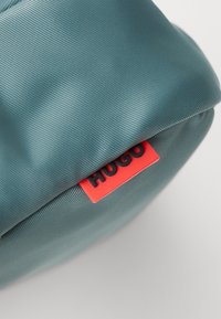 Pungă din material texturat de culoare verde-albastră, cu un eticheta roșie și neagră "HUGO", având o formă rotunjită și o finisare moale.