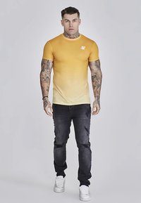 Majica z rumenim prelivom, kratkimi rokavi in okroglim izrezom z logotipom, kombinirana s črnimi raztrganimi kavbojkami in belimi supergami.