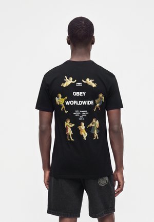 Muž nosí čierne tričko s cherubínmi hrajúcimi na hudobné nástroje okolo textu "Obey Worldwide" a názvov miest vytlačených na chrbte.