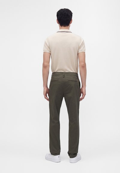 Homme debout, tourné de dos, portant un polo beige à manches courtes, un pantalon vert foncé et des baskets blanches sur un fond uni.