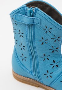 Blå läderankelboot med utskurna blommönster, sidodragkedja och kontrasterande sömmar. Lätt gummisula med texturerad yta.