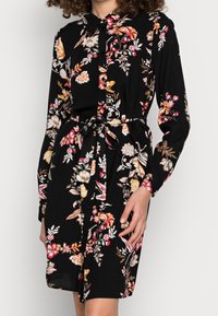 Czarna sukienka koszulowa z długim rękawem, kolorowy kwiatowy nadruk, zapięcie na guziki, związany pasek w talii i kołnierzyk, noszona przez stojącą osobę.